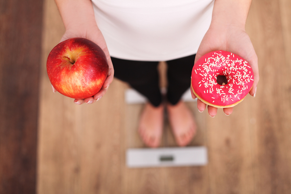 dieta kobieta wazaca sie na wadze w jednej dloni trzyma jablko a w drugiej paczek